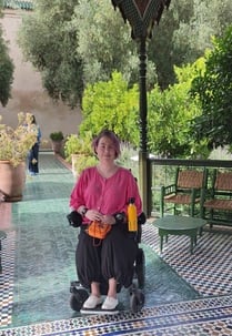 Claire in her electric wheelchair in a beautiful Moroccan garden. She is a pink-haired 40-something white woman wearing a bright pink blouse and black culottes. She is on a path made of intricate blue and green mosaic tiles and surrounded by trees and bushes. Wooden furniture is in the background