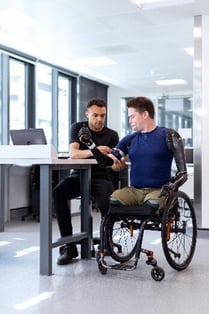 A person in a wheelchair who has had both arms and both legs amputated. He is wearing two prosthetic arms.  A man sat next to him is fitting a device on his bicep.