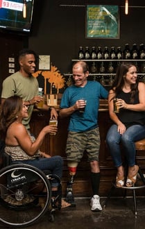 A group of men and women of different ages and races enjoying a drink in a bar. One woman is using a wheelchair. One man has a prosthetic leg.