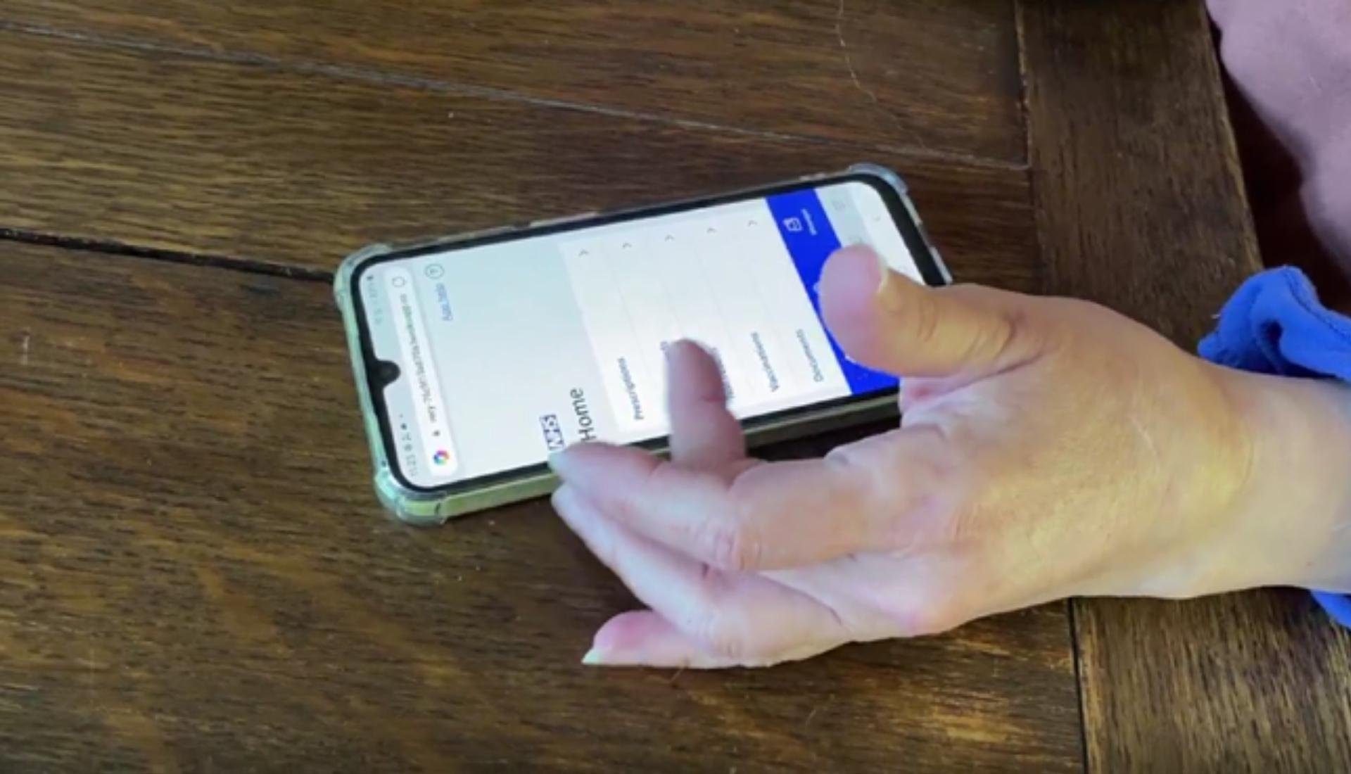 A smartphone rests on the desk as a hand scrolls through the newly designed NHS App homepage.