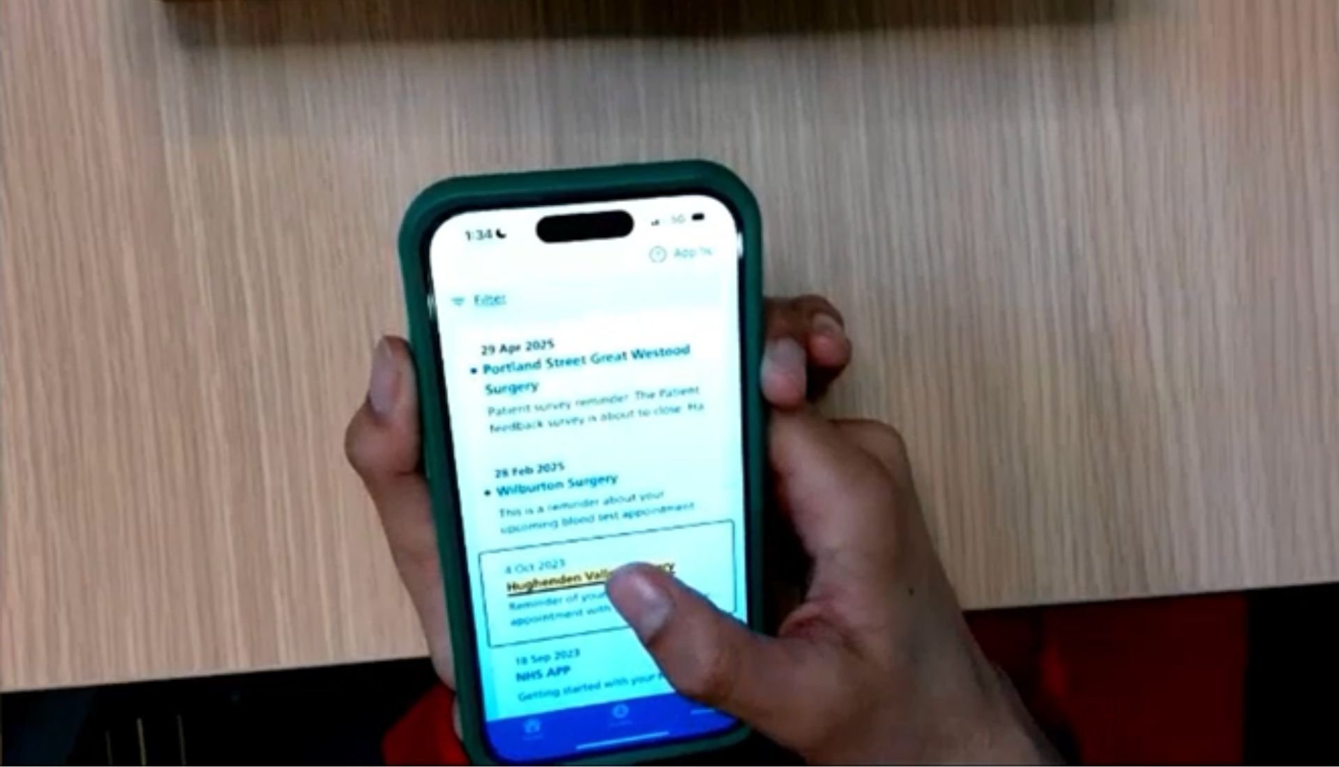 A person holding and clicking the phone with her right thumb. The screen is showing the NHS App messages and the person is using the VoiceOver.